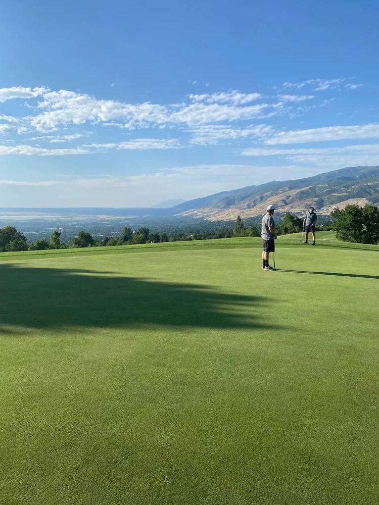 Two golfers on a green at Bountiful Ridge, overlooking the valley and mountain slopes.