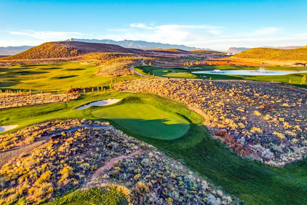 An elevated view of a green beside rocky desert ground, with fairways and a pond beyond.