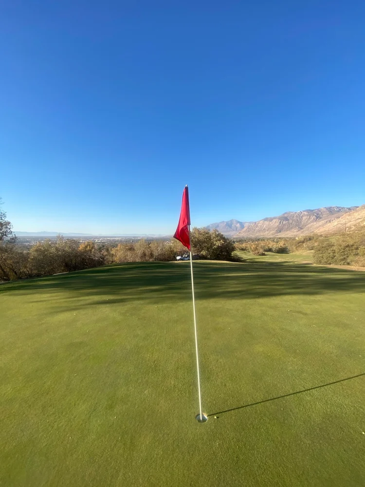 A red flag on a green at Mount Ogden Golf Course, overlooking the valley with mountains to the side.