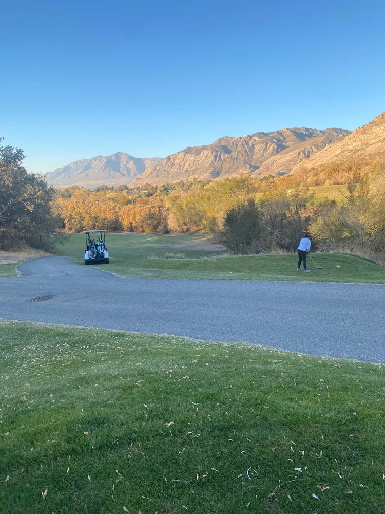 A golfer on a tee beside a cart path at Mount Ogden Golf Course, with trees and rocky mountain slopes beyond.