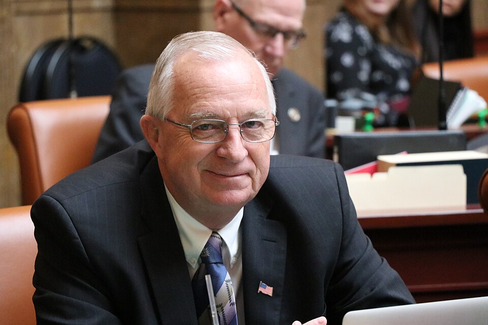Carl Albrecht, wearing glasses and a dark suit, sits at a desk in the Utah House chamber.