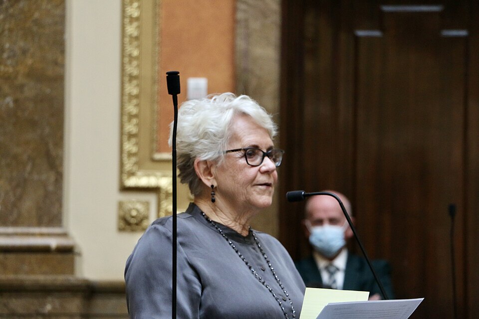 Carol Moss speaking at a microphone while holding papers.