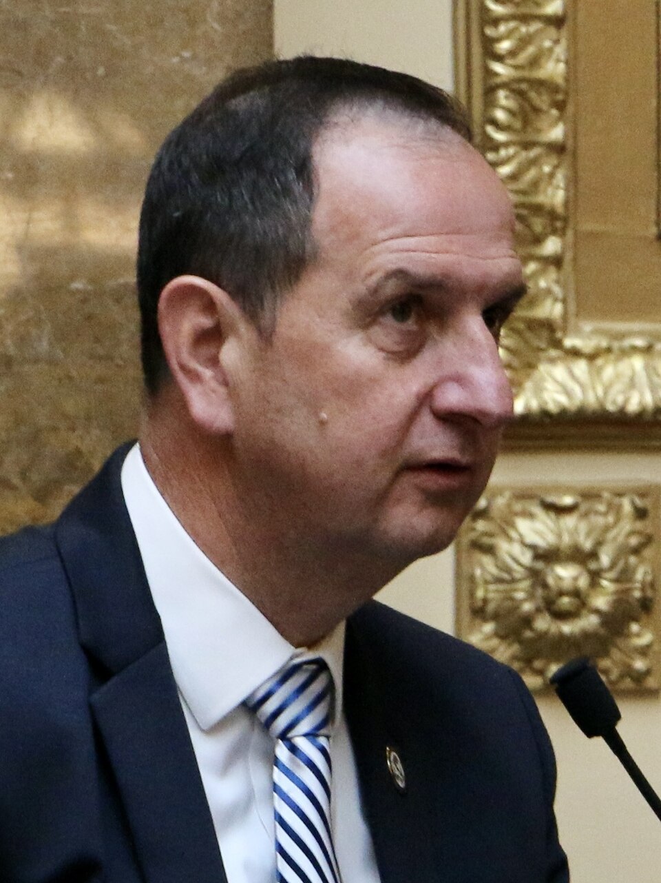 Norm Thurston, wearing a dark suit and striped tie, sits beside a microphone.