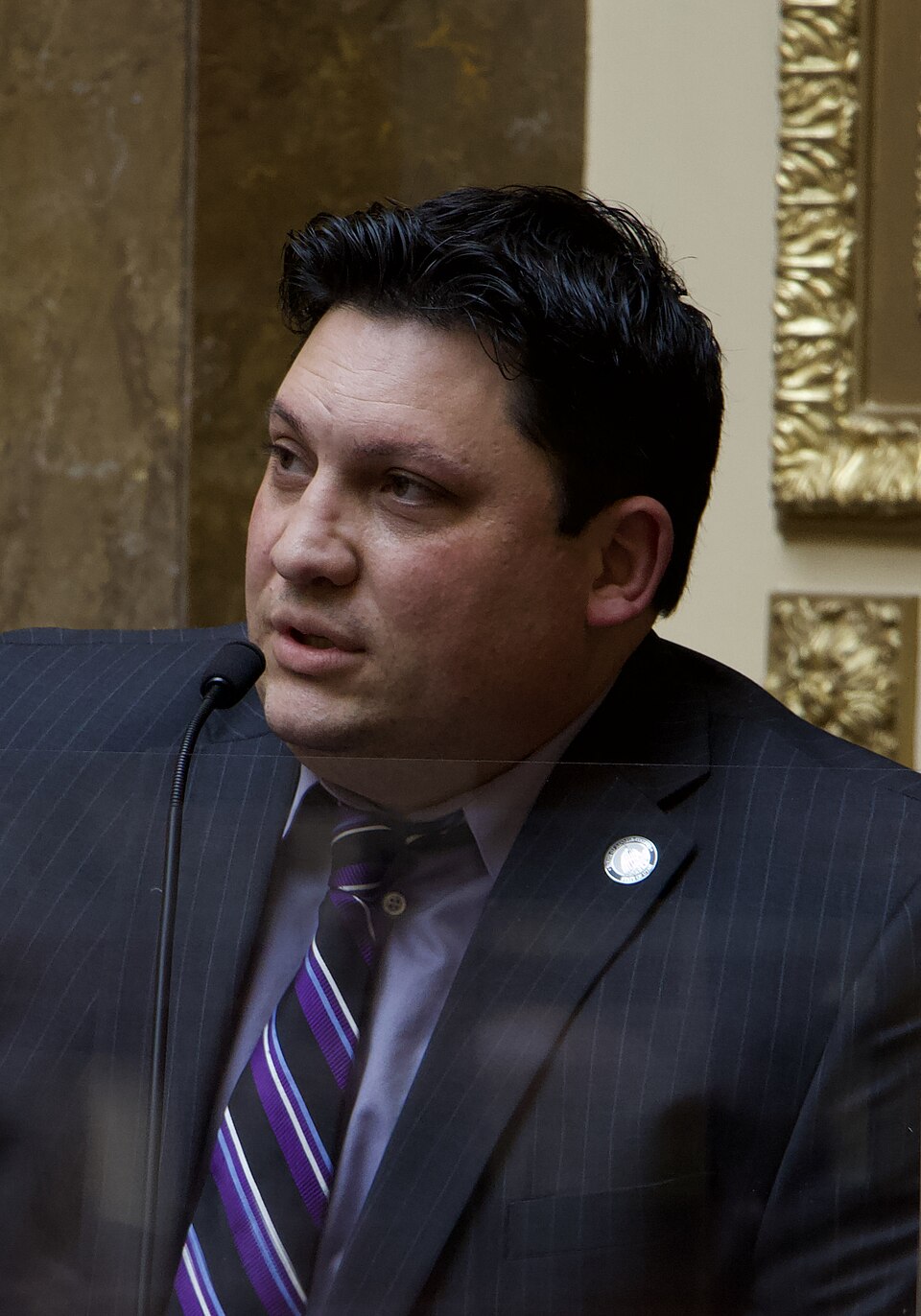 Ryan Wilcox in a dark suit and striped tie speaks into a microphone.