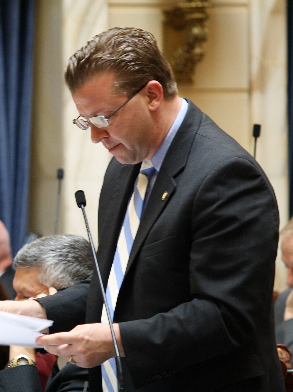 Todd Weiler standing at a chamber microphone reading papers.